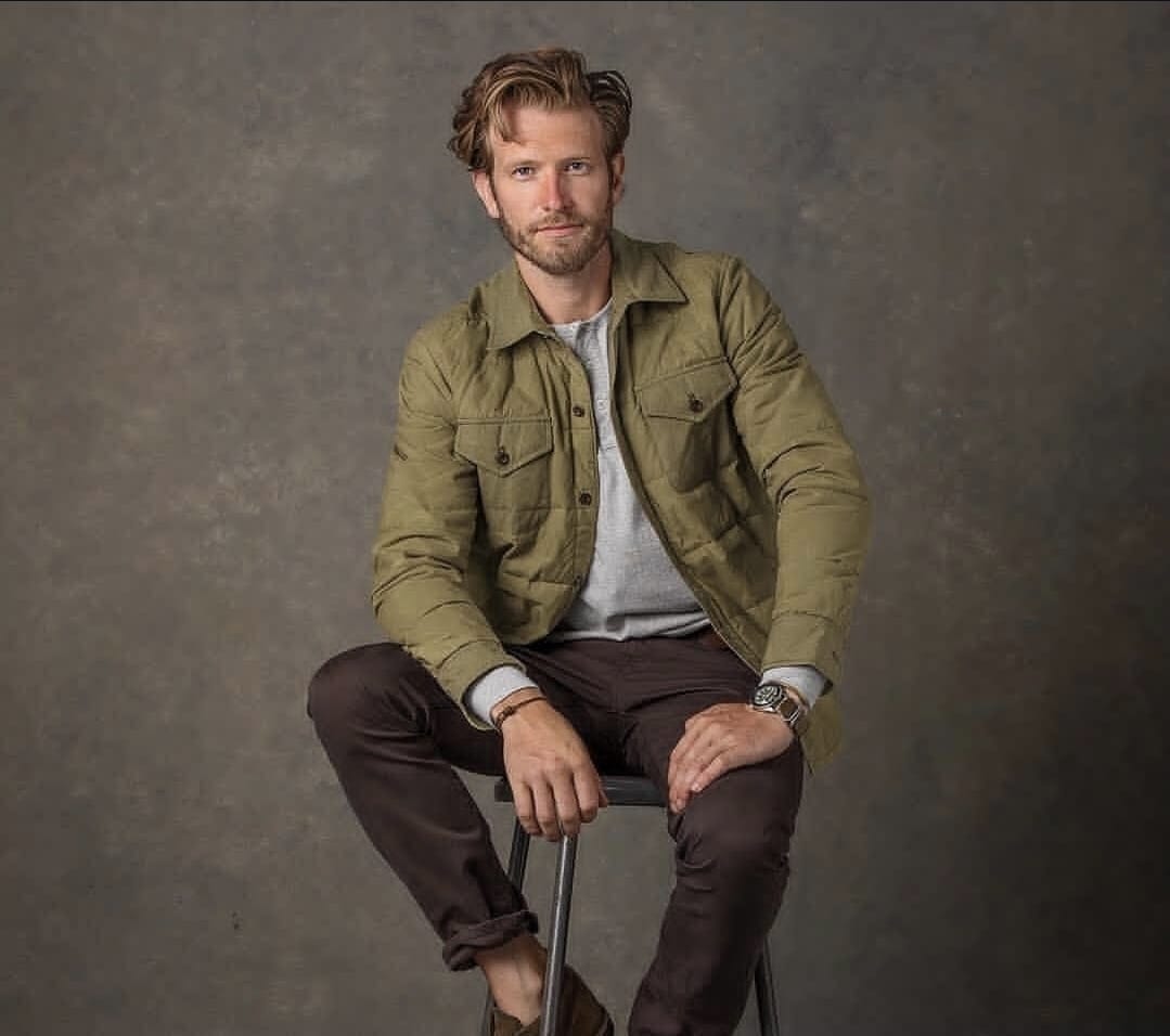 Man in olive quilted jacket seated on stool